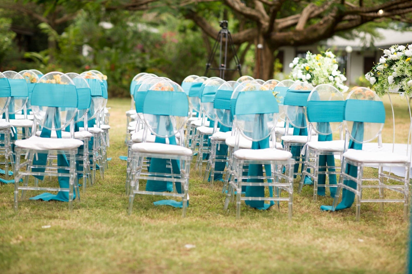Rows of clear chiavari chairs tied with teal ribbons and flowers, ready to welcome guests