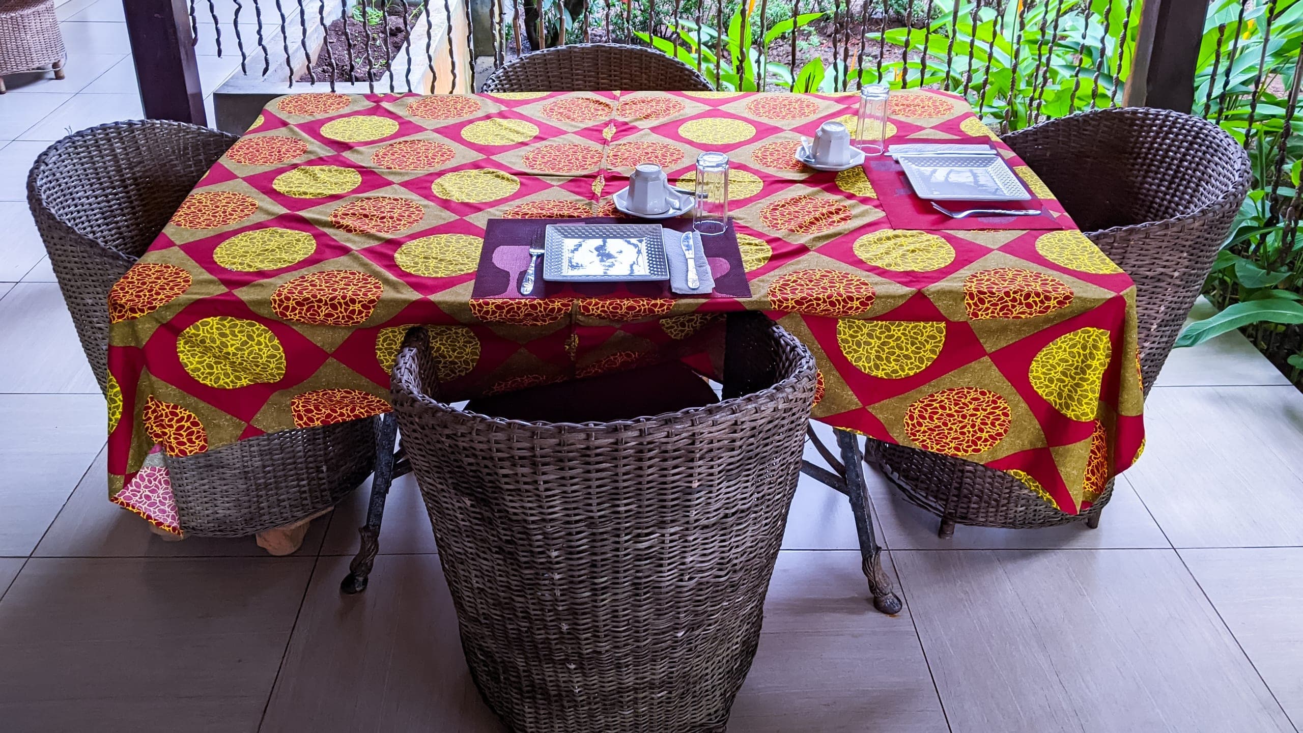 A table dressed for breakfast in a bold African-print cloth, wicker chairs and a lush garden beyond
